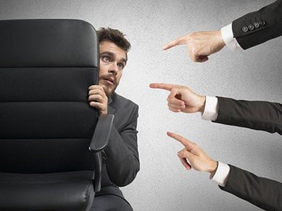 Ein Mann versteckt sich hinter einem Stuhl, während mehrere Hände auf ihn zeigen.